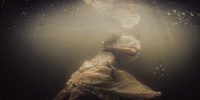 A imagem é do interior de um lago. Há uma mulher submersa até o pescoço, usando um vestido branco