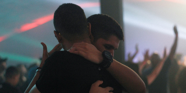 O cenário é uma casa de show e há dois homens se abraçando, um deles visivelmente sentindo-se acolhido.