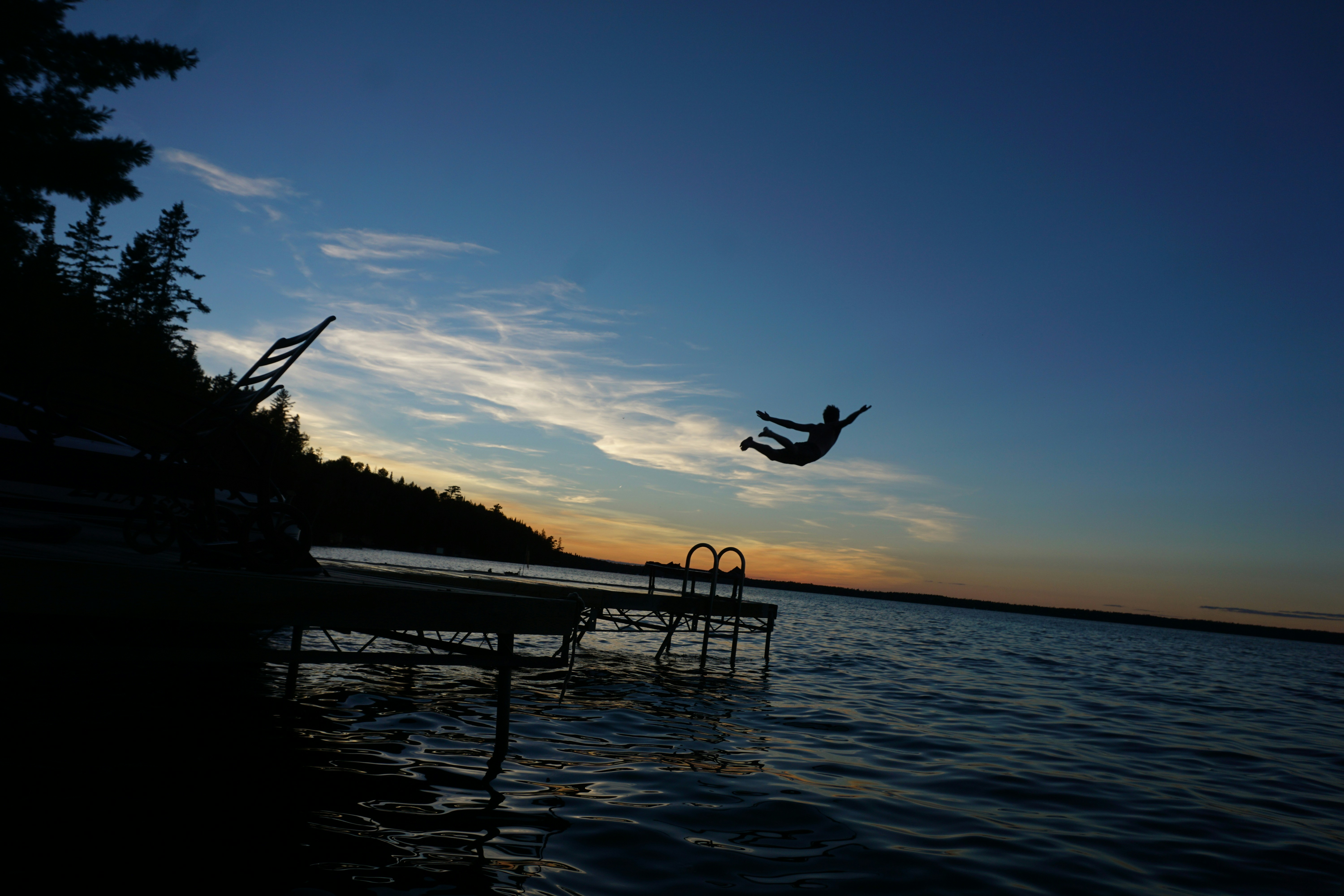 Imagem de um lago, no fim de tarde. Um homem pula do deck com os braços abertos.