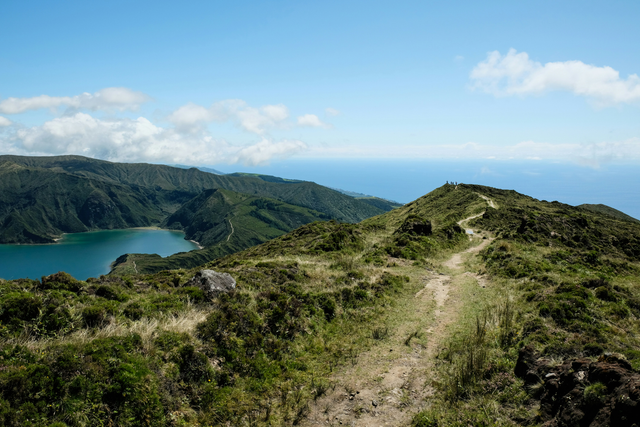 Foto de uma trilha em uma montanha alta, nos Açores. É possível ver um lago e mais ao fundo o Oceano Atlântico.